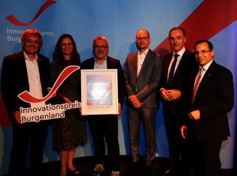 Sechs Personen stehen auf einer Bühne; eine Person in der Mitte hält ein gerahmtes Zertifikat, eine weitere ein Cut-out-Schild mit der Aufschrift 'Innovationspreis Burgenland'. Im Hintergrund ist ein Banner mit dem Logo zu sehen.
