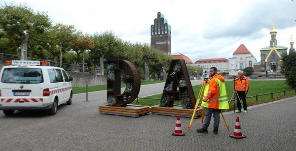 Straßenszene: Zwei Vermessungsarbeiter in Warnwesten stehen neben großen Metallbuchstaben "DA", die auf Holzpaletten liegen. Ein Vermessungsfahrzeug mit der Aufschrift Vermessung steht daneben; Kegel markieren die Arbeitsfläche. Im Hintergrund sind Bäume, eine Kirche und weitere Gebäude zu sehen.