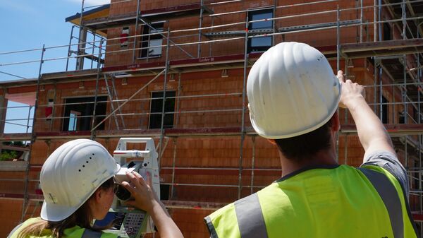 Zwei Bauarbeiter mit weißen Helmen und neon-gelben Warnwesten stehen vor einem im Rohbau befindlichen Holzgebäude mit Gerüst; einer blickt durch ein Messgerät, der andere zeigt in Richtung des Bauwerks.