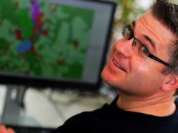 Ein Mann mit Brille lächelt in die Kamera; im Hintergrund sieht man einen Computerbildschirm mit einer farbigen Karten- bzw. Wetteransicht.