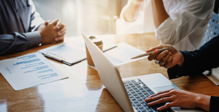 Trois professionnels collaborent autour d'un ordinateur portable sur une table en bois; des documents et des graphiques sont posés devant eux.