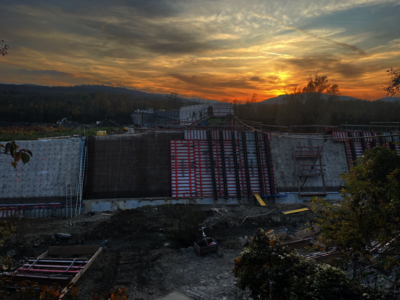 Construction site at sunset: a long concrete wall under formwork panels, red-and-white striped supports, and scattered materials in the foreground, with hills and trees in the distance.