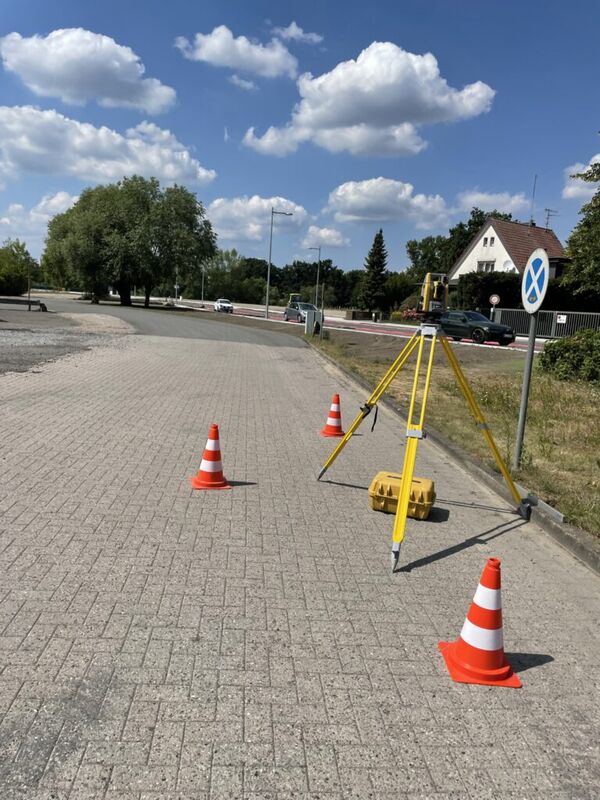 Ein Vermessungsgerät auf Stativ steht am Straßenrand, umgeben von orangefarbigen Verkehrsleitkegeln. Hinter der Baustelle sieht man Straße, Bäume und ein Haus; der Himmel ist blau mit Wolken.