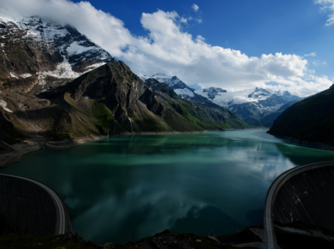Türkisfarbener Bergsee in einem engen Alpental, umgeben von schroffen, schneebedeckten Gipfeln. Die Wasseroberfläche spiegelt Himmel und Wolken wider; im Vordergrund sind Bogenstrukturen eines Damms sichtbar.