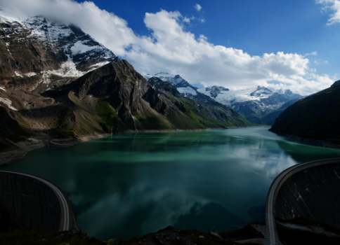 Türkisfarbener Bergsee in einem engen Alpental, umgeben von schroffen, schneebedeckten Gipfeln. Die Wasseroberfläche spiegelt Himmel und Wolken wider; im Vordergrund sind Bogenstrukturen eines Damms sichtbar.