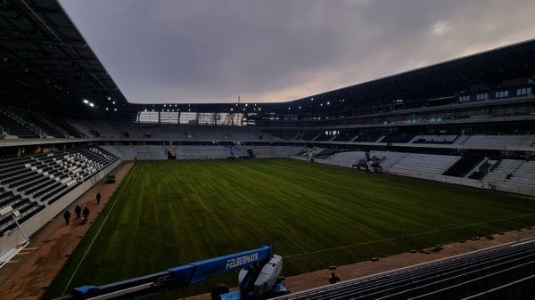 Leeres Fußballstadion mit grünem Rasen, umlaufenden Tribünen und grauen Wolken; wenige Personen stehen an der Seitenlinie.