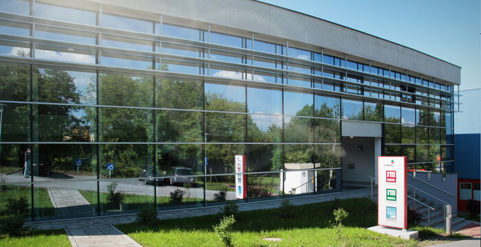 Façade d’un bâtiment moderne avec de grandes baies vitrées reflétant les arbres. Entrée sur la droite avec escaliers et signalétique colorée à proximité. Parking et accès devant le bâtiment.
