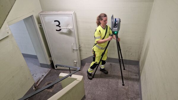 Eine Person in gelber Sicherheitskleidung bedient ein Vermessungsgerät auf einem Stativ in einem Treppenhaus.