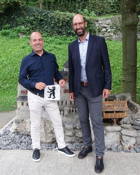Zwei Männer stehen draußen vor einer Steinmauer, umgeben von Gras, und lächeln in die Kamera. Der Mann links trägt ein dunkelblaues Hemd und weiße Hosen und hält ein Schild mit einem schwarzen Löwen-Emblem. Der Mann rechts trägt Blazer, helles Hemd und graue Hosen.
