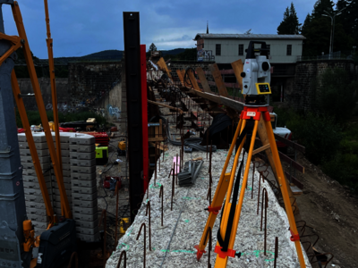 Construction site with a yellow-orange tripod-mounted surveying instrument on a gravel path. Rebar stakes line the sides, outlining a foundation. In the background are building materials, a partially built wall, and a distant structure under a cloudy sky.