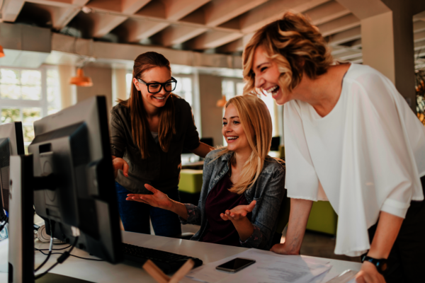 Drei Kolleginnen stehen um einen Computerbildschirm in einem offenen Büro. Die sitzende blonde Frau erklärt etwas, die beiden anderen lachen und gestikulieren. Auf dem Tisch liegen Unterlagen und ein Smartphone; Licht fällt durch große Fenster.