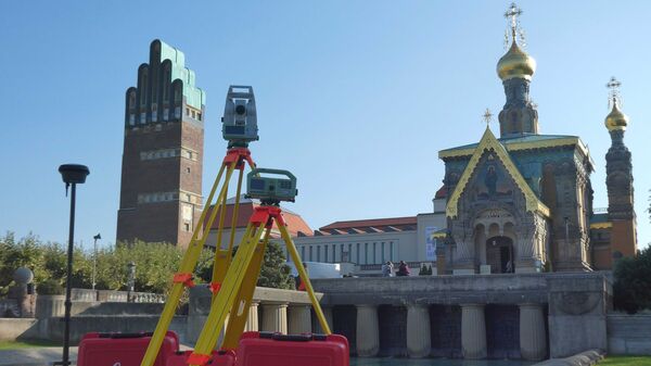 Vordergrund: Vermessungsgerät auf einem Dreibein-Stativ. Hintergrund: orthodoxe Kirche mit goldenen Kuppeln vor klarem blauen Himmel.