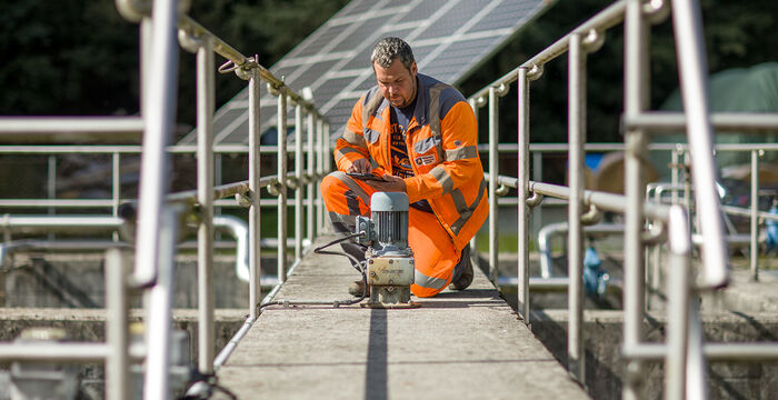 Un ouvrier en combinaison orange haute visibilité est agenouillé sur une passerelle métallique, utilisant un outil électrique posé au centre pour réaliser des travaux de maintenance sur une structure extérieure.