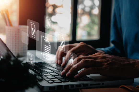 Nahaufnahme von Händen, die auf einer Laptop-Tastatur tippen; darüber schweben transparente Dokumente, die digitale Arbeiten symbolisieren. Hintergrund unscharf mit warmem Fensterlicht.