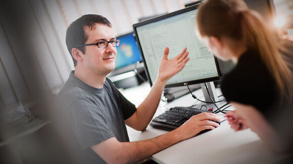 Two coworkers in an office discuss at a desk with computer screens; a man wearing glasses gestures toward the monitor as a woman listens and looks at documents.