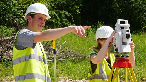 Zwei Bauarbeiter tragen Helme und Warnwesten im Freien. Einer zeigt mit dem Arm, der andere bedient eine Vermessungsstation auf einem Stativ; sie führen eine Vermessung im Grünen durch.
