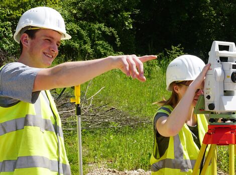Zwei Bauarbeiter tragen Helme und Warnwesten im Freien. Einer zeigt mit dem Arm, der andere bedient eine Vermessungsstation auf einem Stativ; sie führen eine Vermessung im Grünen durch.