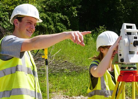 Zwei Bauarbeiter tragen Helme und Warnwesten im Freien. Einer zeigt mit dem Arm, der andere bedient eine Vermessungsstation auf einem Stativ; sie führen eine Vermessung im Grünen durch.