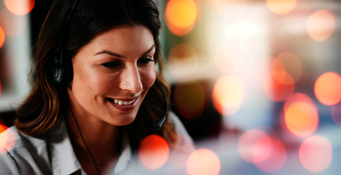 Eine freundliche Frau mit Headset, vermutlich Kundendienstmitarbeiterin, lächelt ins Mikrofon. Der Hintergrund ist unscharf mit bunten Lichterflecken.