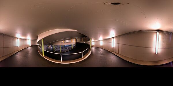 Eine helle Innenpassage in einem Tunnel: In der Mitte steht ein runder Geländerrahmen; der umlaufende Korridor wird von glatten weißen Wänden flankiert und von Wandlampen beleuchtet.