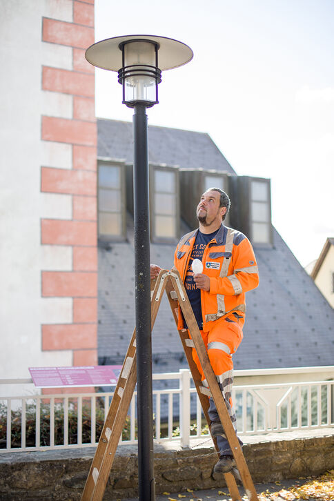 Ein Arbeiter in orange Sicherheitskleidung steht auf einer Leiter und repariert eine Straßenlaterne; dahinter sind Gebäude und ein Geländer sichtbar.
