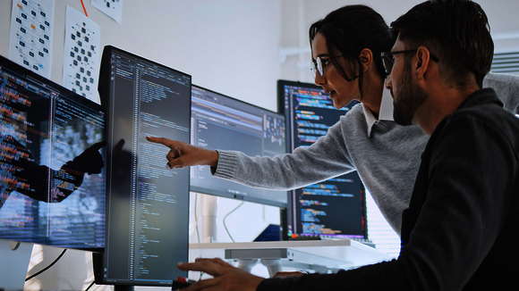 Zwei Entwickler arbeiten gemeinsam an mehreren Monitoren mit Code; eine Person zeigt mit dem Zeigfinger auf eine Codezeile, während der andere zuschaut. Eine helle Arbeitsumgebung im Hintergrund.