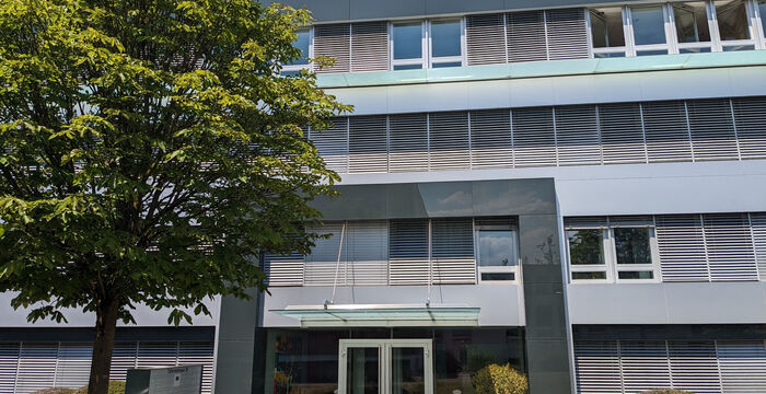Façade d'un bâtiment moderne avec une entrée en verre sous un auvent, flanquée de deux buissons; un grand arbre à gauche et un ciel bleu clair en arrière-plan.
