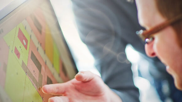 Eine Person mit Brille schaut auf einen Computerbildschirm, der ein farbiges Layout mit quadratischen Blöcken in Pink, Grün und Gelb zeigt; eine Hand zeigt auf das Display.