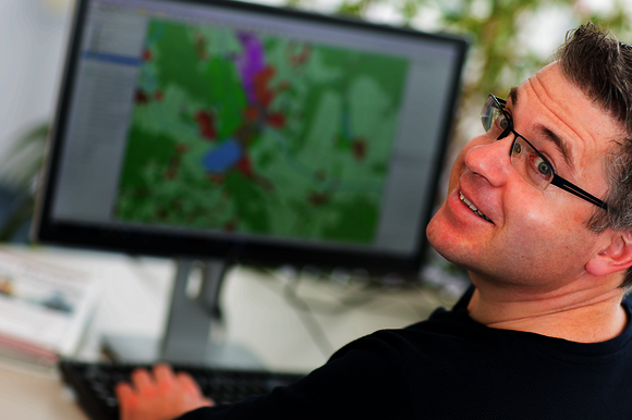 Ein Mann mit Brille sitzt an einem Schreibtisch; er lächelt in die Kamera. Auf dem Monitor im Hintergrund ist eine bunte Karte zu sehen.