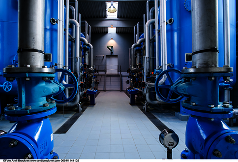 Ansicht einer Industrieanlage mit symmetrisch angeordneten blauen Rohrleitungen, Ventilen und Pumpen. Ein mittlerer Gang führt zu einer Tür im hinteren Bereich, von Deckenleuchten beleuchtet. Die Szene vermittelt eine hochspezialisierte Wartungsumgebung.
