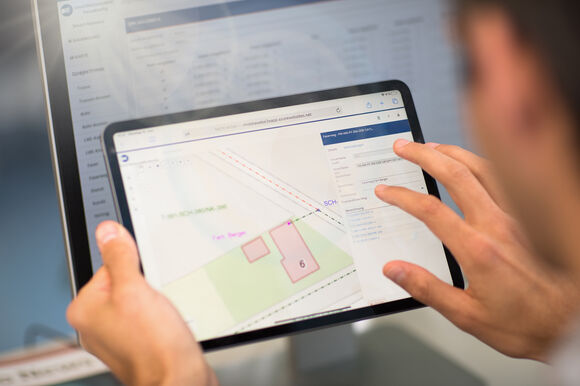 Eine Person hält ein Tablet; darauf ist eine Kartenansicht mit farbigen Feldern, Linien und nummerierten Bereichen zu sehen.
