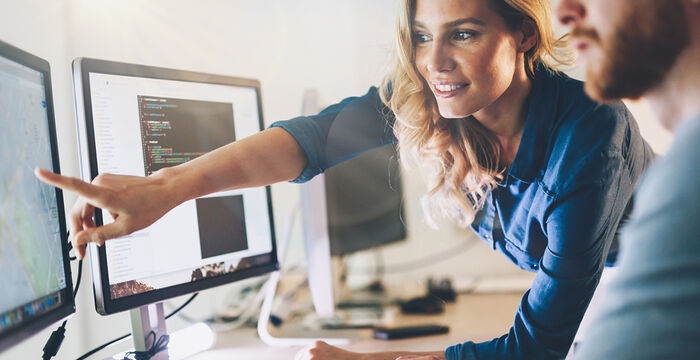 Zwei Personen in einem Büro arbeiten an Computern; eine Frau zeigt auf den Monitor, auf dem Programmiercode zu sehen ist, während der Mann daneben zusieht.