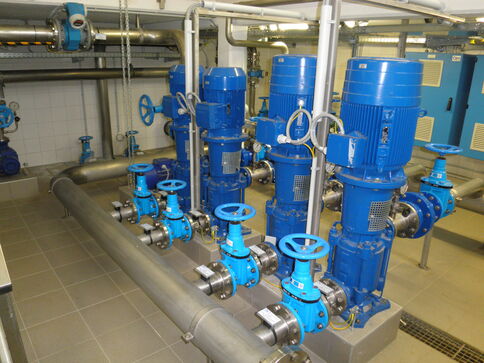 Industriepumpenanlage: Mehrere blaue Pumpenmotoren mit Ventilen und Rohrleitungen in einem Technikraum; sichtbare Handrääder, Steuereinheiten und Verdrahtung.