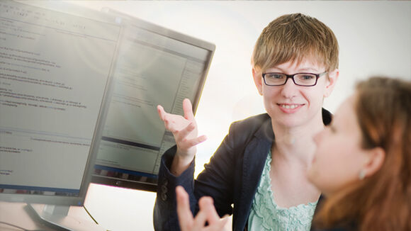 Two colleagues discuss code at a computer workstation: a woman with short hair and glasses smiles while gesturing toward a monitor displaying lines of code; another person sits nearby, slightly out of focus.