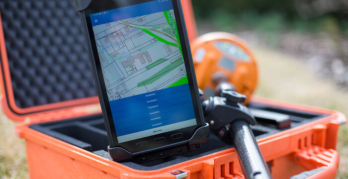 Smartphone mit Kartenanzeige in einem robusten orangefarbenen Koffer im Freien. Auf dem Display ist eine Kartenansicht mit grünen Linien zu sehen, während ein orangefarbenes Messgerät im Hintergrund liegt.