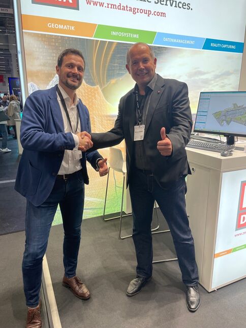 Zwei Männer stehen auf einem Messestand und schütteln sich die Hände, lächeln in die Kamera. Einer zeigt Daumen hoch. Im Hintergrund sind Monitor und Werbebanner des Ausstellers zu sehen.