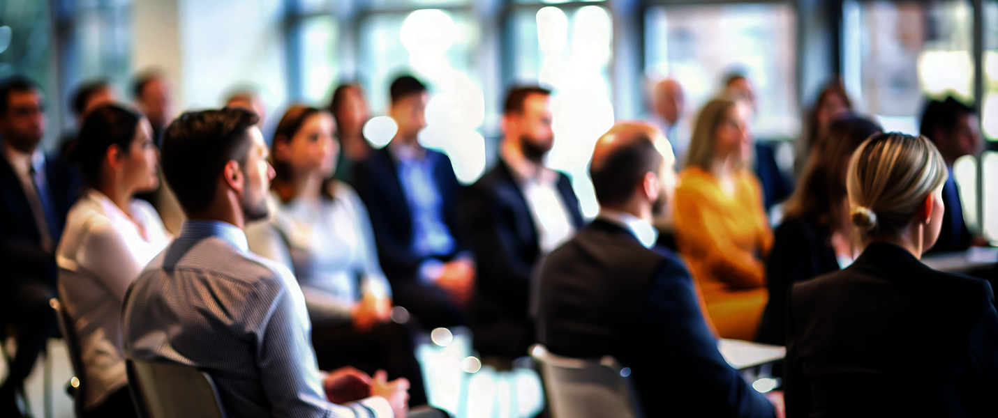 Eine Gruppe von Berufstätigen in Business-Kleidung sitzt in einem Konferenzraum und hört einer Präsentation zu.