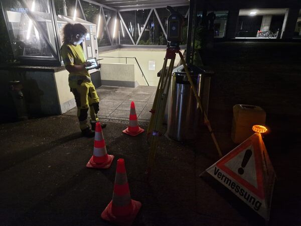 Nachtaufnahme: Eine Person in gelber Warnschutzkleidung steht vor einem Gebäudeeingang, hält ein Tablet und bedient ein Vermessungsgerät auf einem Stativ. Vor ihr stehen Verkehrsleitkegel, ein Warnschild "Vermessung" und eine Sicherheitsbeleuchtung; Bereich wirkt abgesperrt.