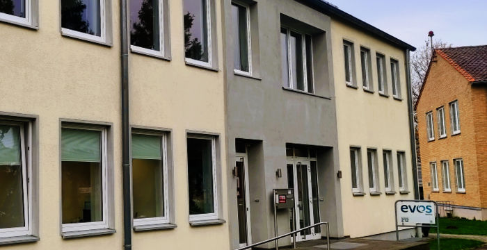 Zweistöckiges Gebäude mit gelblicher Fassade und vielen rechteckigen Fenstern. Zentraler Eingang mit Treppe und Rampe sowie Metallhandläufen. Vor dem Gebäude befindet sich ein EVOS-Schild und eine kleine Grünfläche.