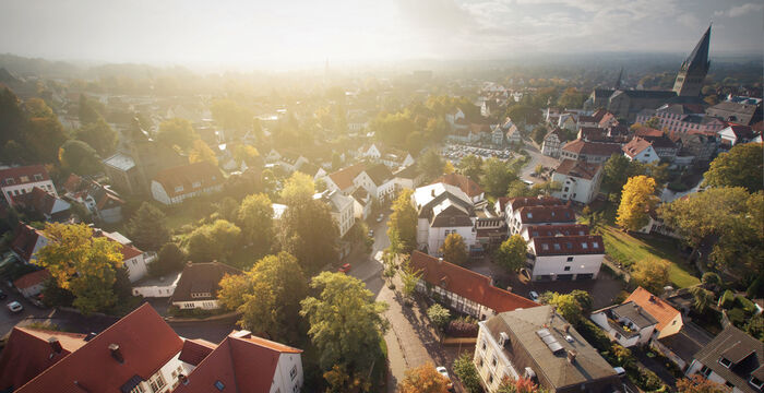 Vue aérienne d'une petite ville au lever du soleil: maisons aux toits rouges et gris, rues étroites et arbres généreux; au loin, une église avec clocher domine le paysage.